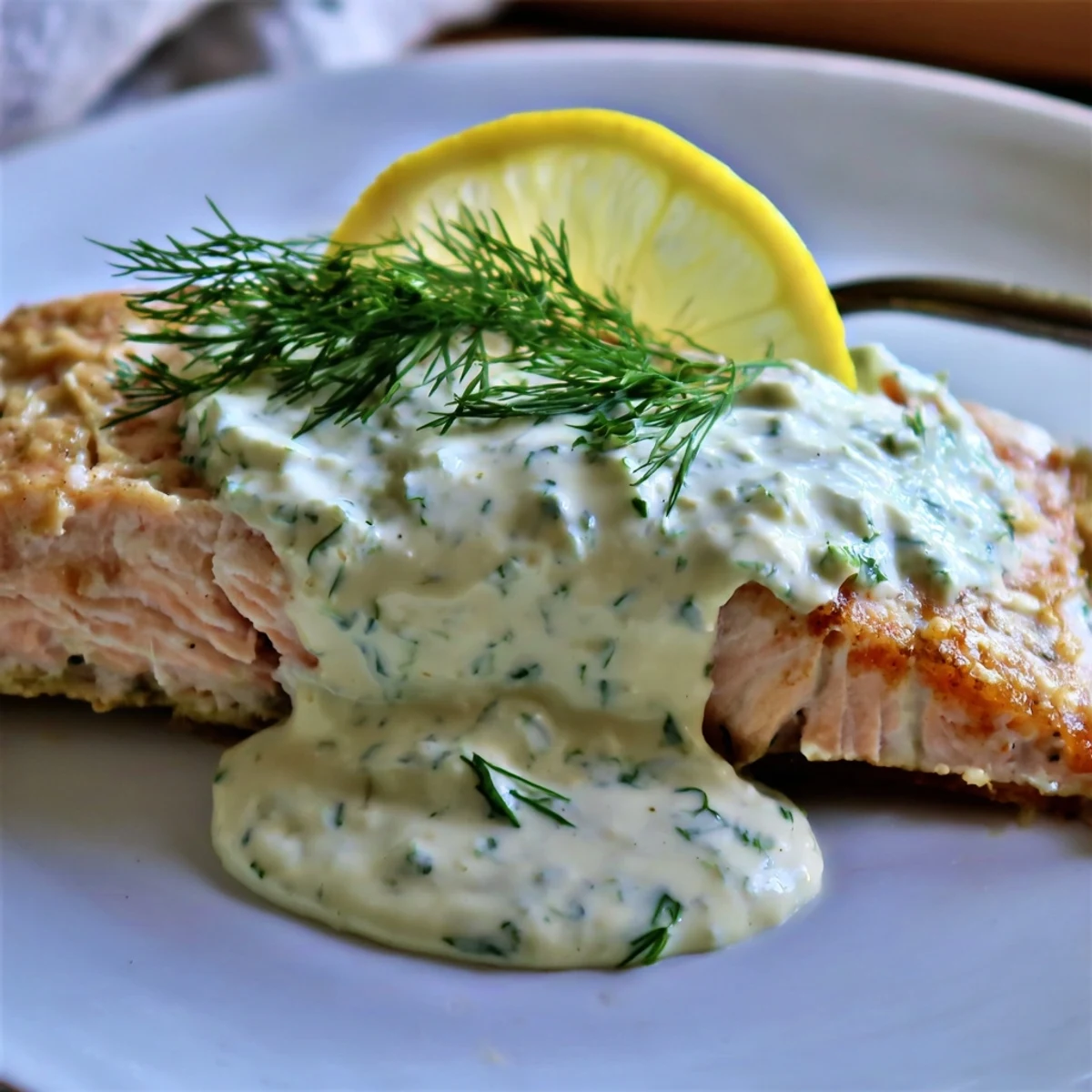 Perfectly baked salmon fillet flaking easily, topped with a creamy, pale green dill sauce and fresh lemon slices.