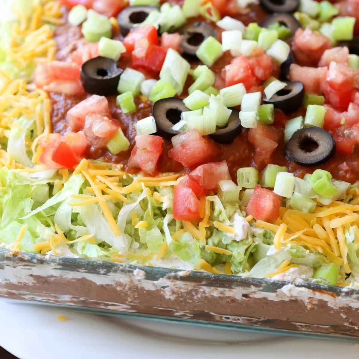 Close-up of Seven Layer Taco Dip with lettuce, tomatoes, and olives alongside tortilla chips, ready to serve.