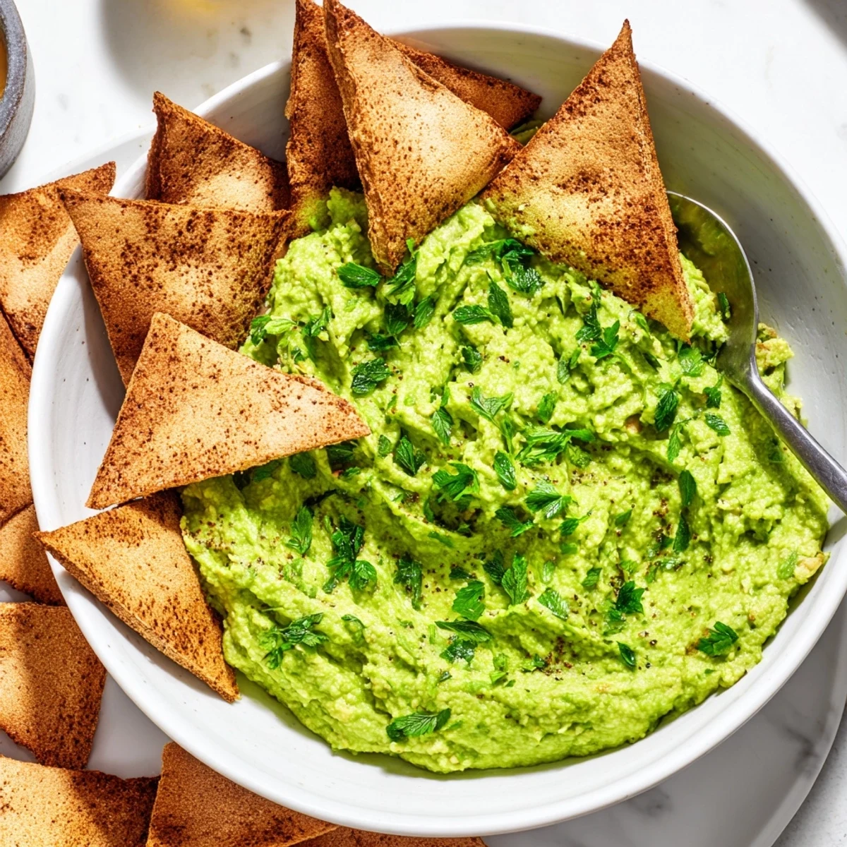 Fresh Green Avocado Hummus with Pita Chips is shown in a white bowl, garnished with herbs and olive oil.
