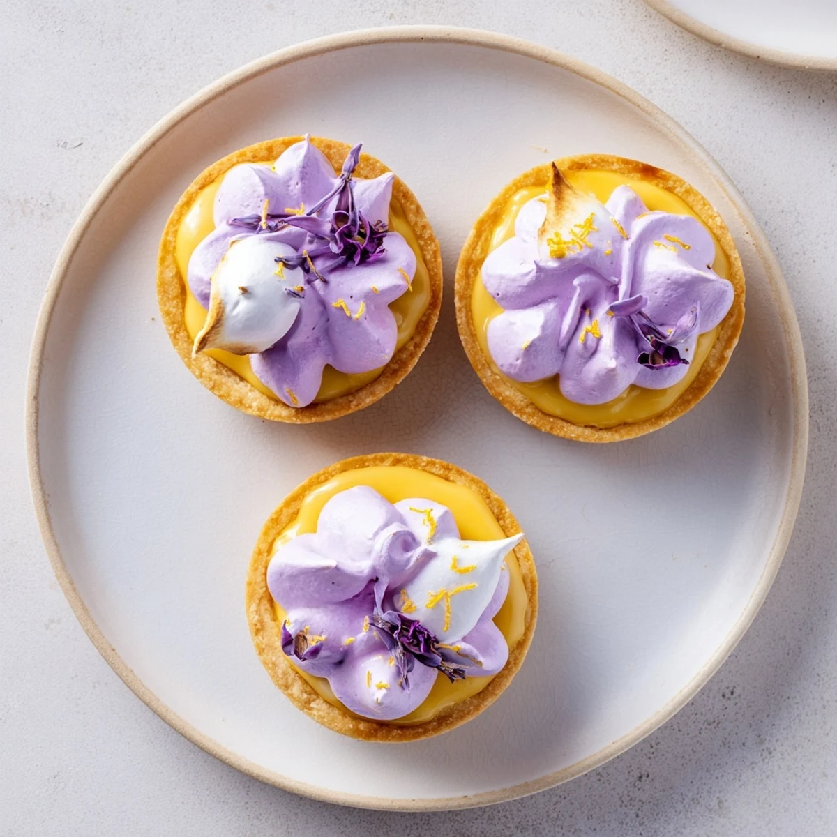 Golden mini lemon tart featuring lilac meringue garnished with fresh edible purple blossoms on white plate