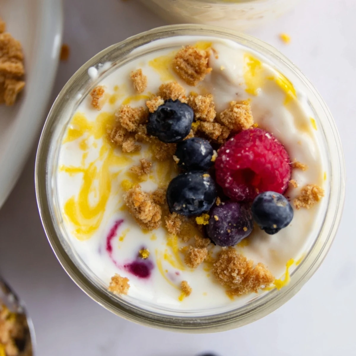 Glass jar layered with zesty lemon cheesecake overnight oats topped with fresh blueberries and graham cracker crumbs