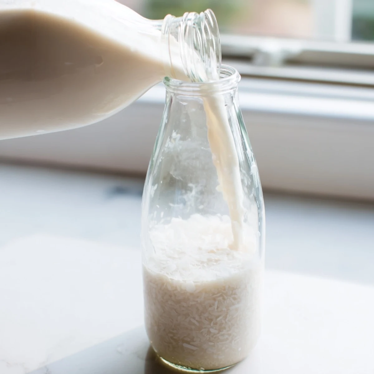 Creamy white homemade coconut milk poured into clear glass jar on wooden counter