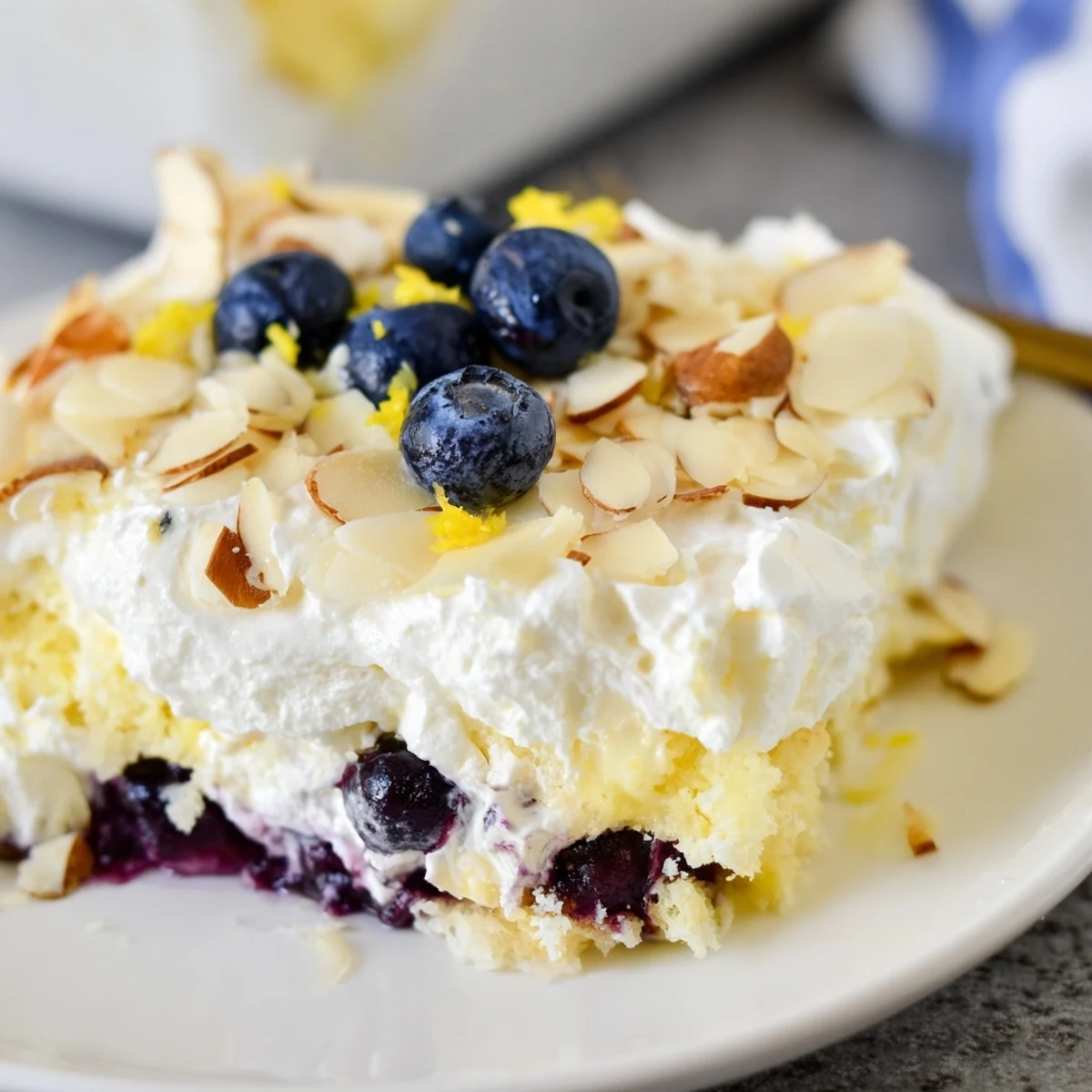 Golden slice of lemon blueberry poke cake topped with fluffy whipped cream and toasted almonds